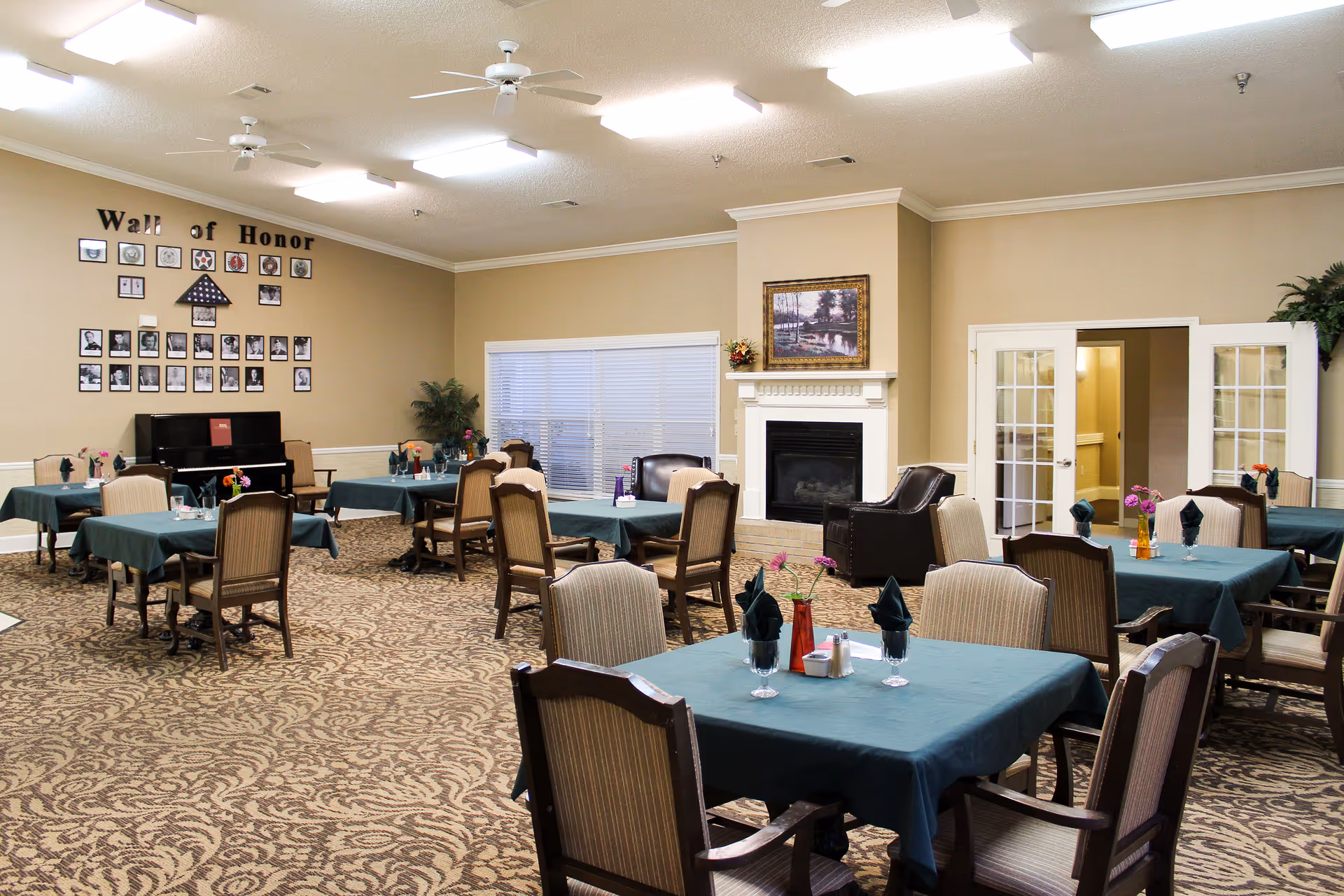 Spacious dining room with several tables set with green tablecloths, chairs, a fireplace, and a 'Wall of Honor' photo display.
