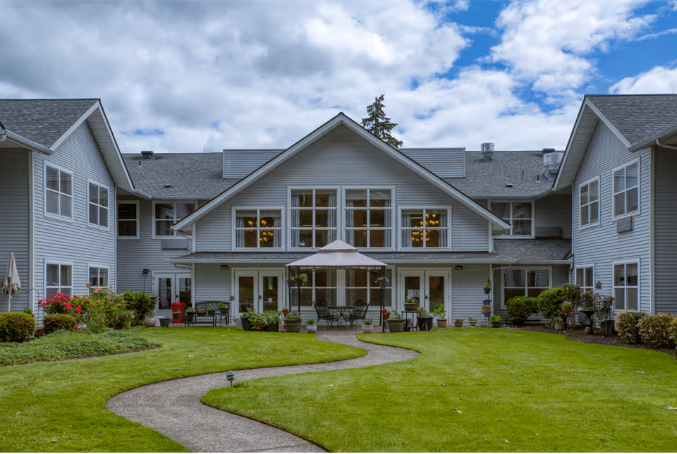 Exterior view of Homewood Assisted Living facility showing a two-story building with gray siding and multiple windows. There is a well-maintained green lawn with a curved concrete pathway leading to a patio area with outdoor tables, chairs, and a large umbrella. The sky is partly cloudy.