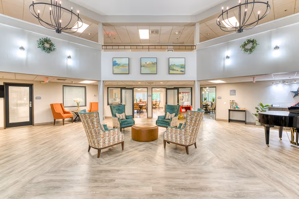 A spacious senior living facility common area with a circular seating arrangement consisting of four patterned armchairs and two teal armchairs around a round brown ottoman. The room has light wood flooring, white walls, and two large chandeliers hanging from the ceiling. There are three landscape paintings on the far wall above glass doors leading to a dining area. Additional seating with orange chairs and a small table is visible on the left, and a black grand piano is on the right side of the room.