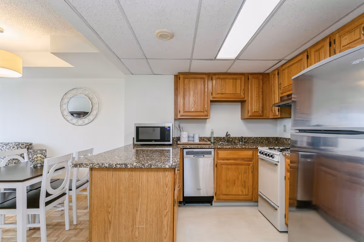 A kitchen area in a senior living facility featuring wooden cabinets, a granite countertop, a stainless steel refrigerator, a dishwasher, a microwave, and a stove. Adjacent to the kitchen is a dining area with a dark wooden table and white chairs. A round decorative mirror hangs on the white wall.