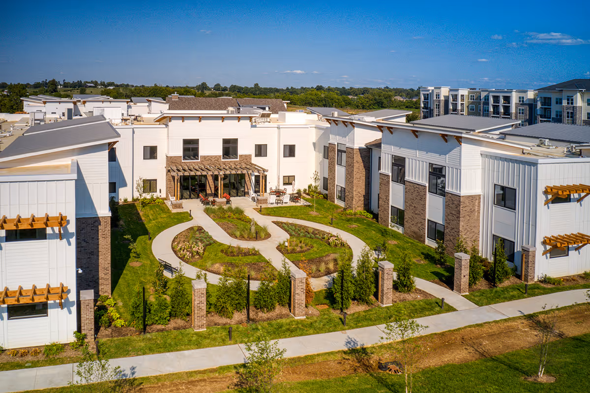 Aerial view of Preston Greens Senior Living facility showing a modern two-story building with white and brick exterior walls. The building surrounds a landscaped courtyard with winding concrete pathways, green grass, shrubs, and outdoor seating areas under pergolas. The sky is clear and blue.