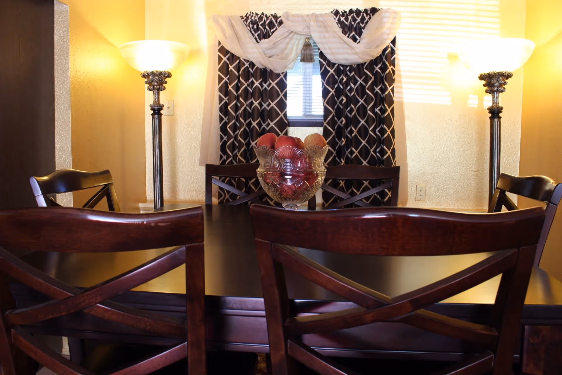 A dining room with a dark wooden table and matching chairs. On the table is a glass bowl filled with red apples. Behind the table is a window with black and white patterned curtains and a sheer white swag valance. Two tall floor lamps with frosted glass shades are positioned on either side of the window, casting warm light on the beige walls.