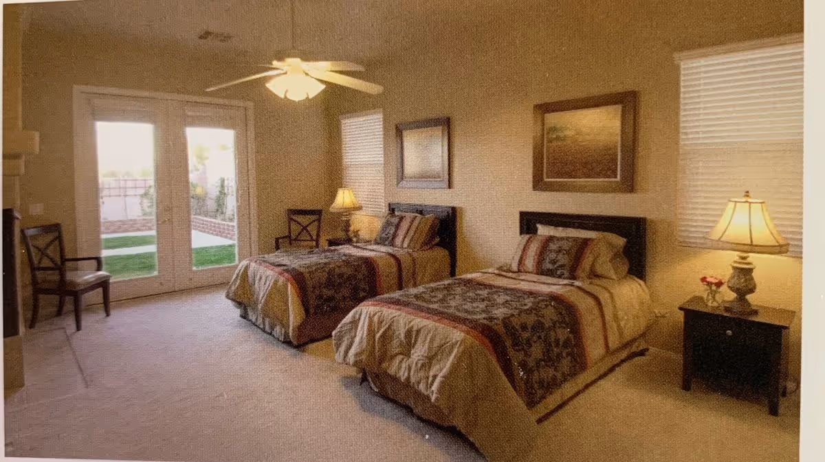 A bright bedroom with two twin beds, nightstands and lamps, a ceiling fan, framed art, chairs, and French doors opening to a yard.