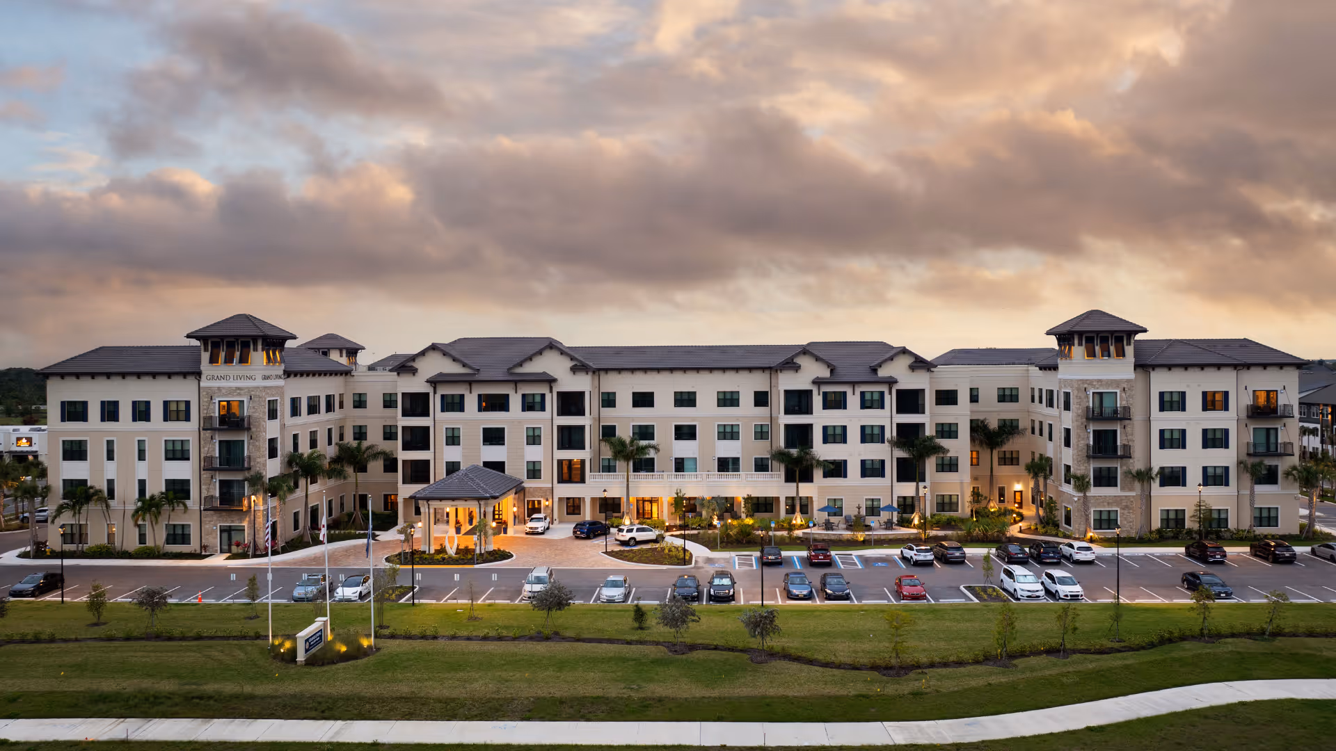 Exterior view of a large, modern senior living facility named Grand Living at Wellen Park during sunset, with a spacious parking lot in front and well-maintained landscaping including palm trees and grass.