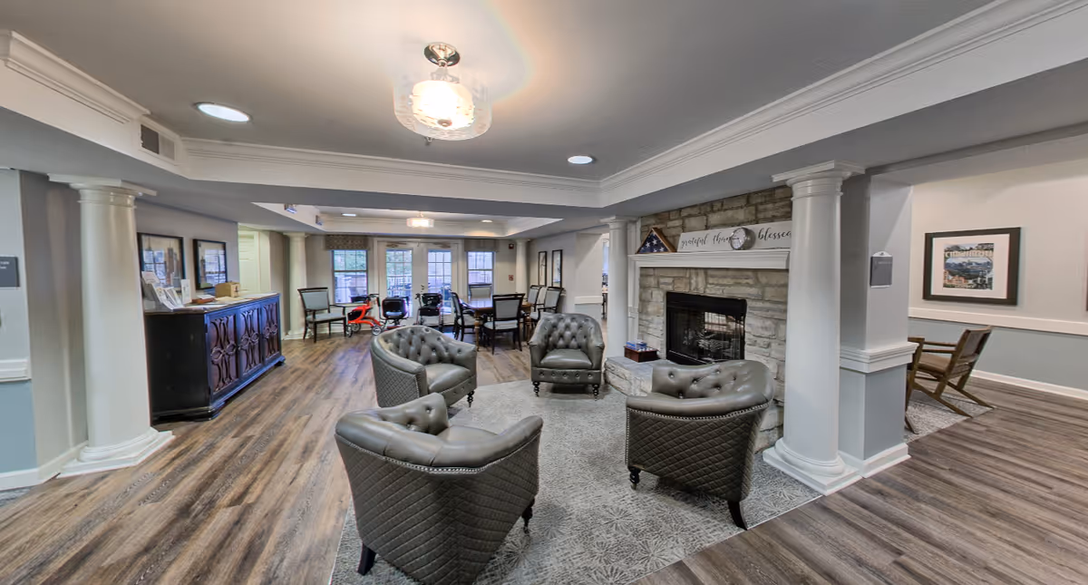 A spacious senior living common room with tufted leather chairs arranged around a fireplace, columns, and a dining area in the background.