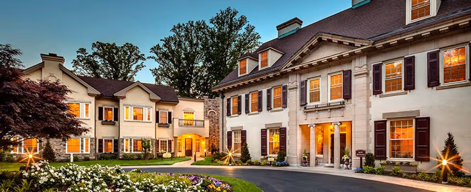 Exterior view of a large, elegant senior living facility building at dusk with warm lights glowing from the windows, surrounded by landscaped gardens and trees.