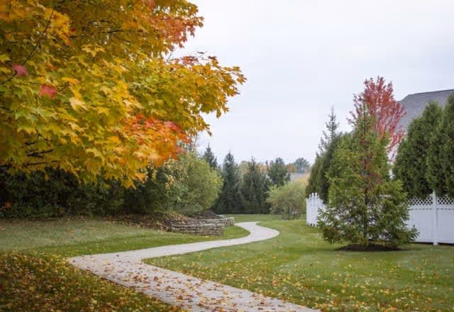 A winding concrete pathway through a grassy area with trees showing autumn colors, including yellow and red leaves. There is a white fence on the right side and a stone retaining wall on the left side, with more trees and shrubs in the background under a cloudy sky.
