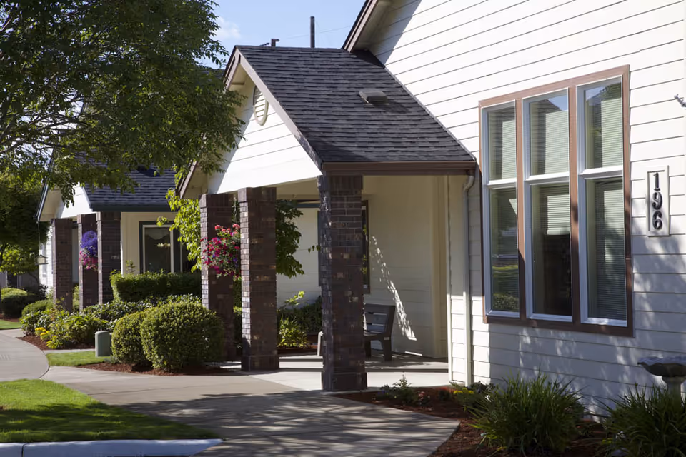 Exterior view of a senior living facility with white siding and dark shingled roofs. The building features covered walkways supported by brick pillars, with hanging flower baskets and well-maintained landscaping including bushes and trees. A bench is visible under one of the covered areas.