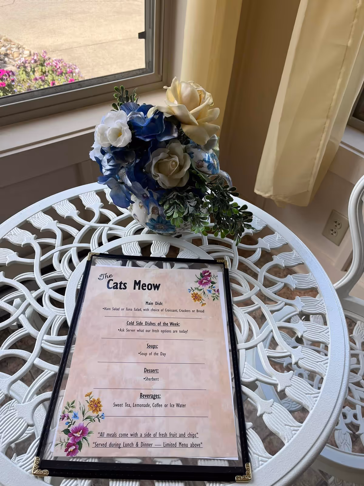 A white ornate metal table with a floral arrangement in a blue and white vase and a menu titled 'The Cats Meow' placed on the table. The table is near a window with beige curtains, and part of a white chair is visible.
