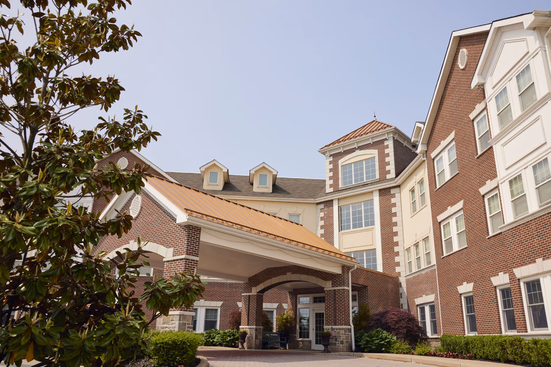 Exterior view of a senior living facility building with red brick walls, white trim, and a covered entrance with a copper-colored roof. There are windows on multiple floors and landscaping with bushes and a tree in the foreground under a clear sky.