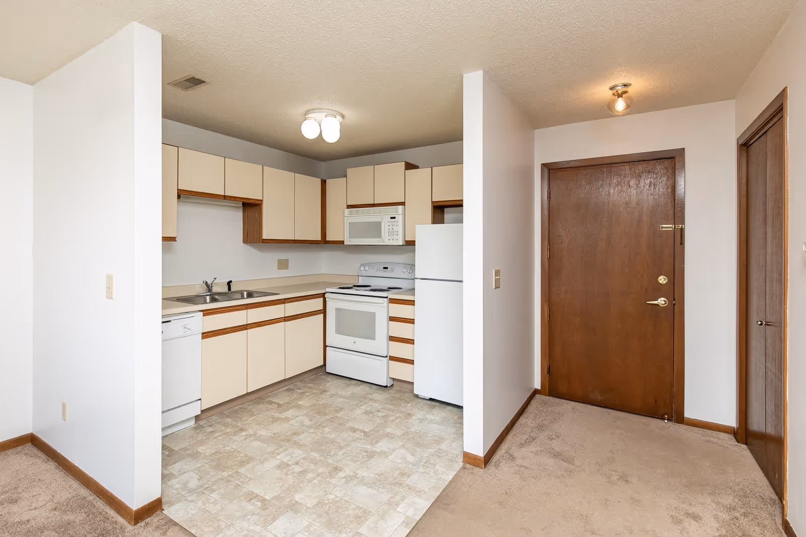 Interior view of a senior apartment kitchen area with beige cabinets, white appliances including a refrigerator, stove, microwave, and dishwasher. The kitchen floor is tiled, and adjacent to it is a carpeted entryway with a wooden front door and a closet door.
