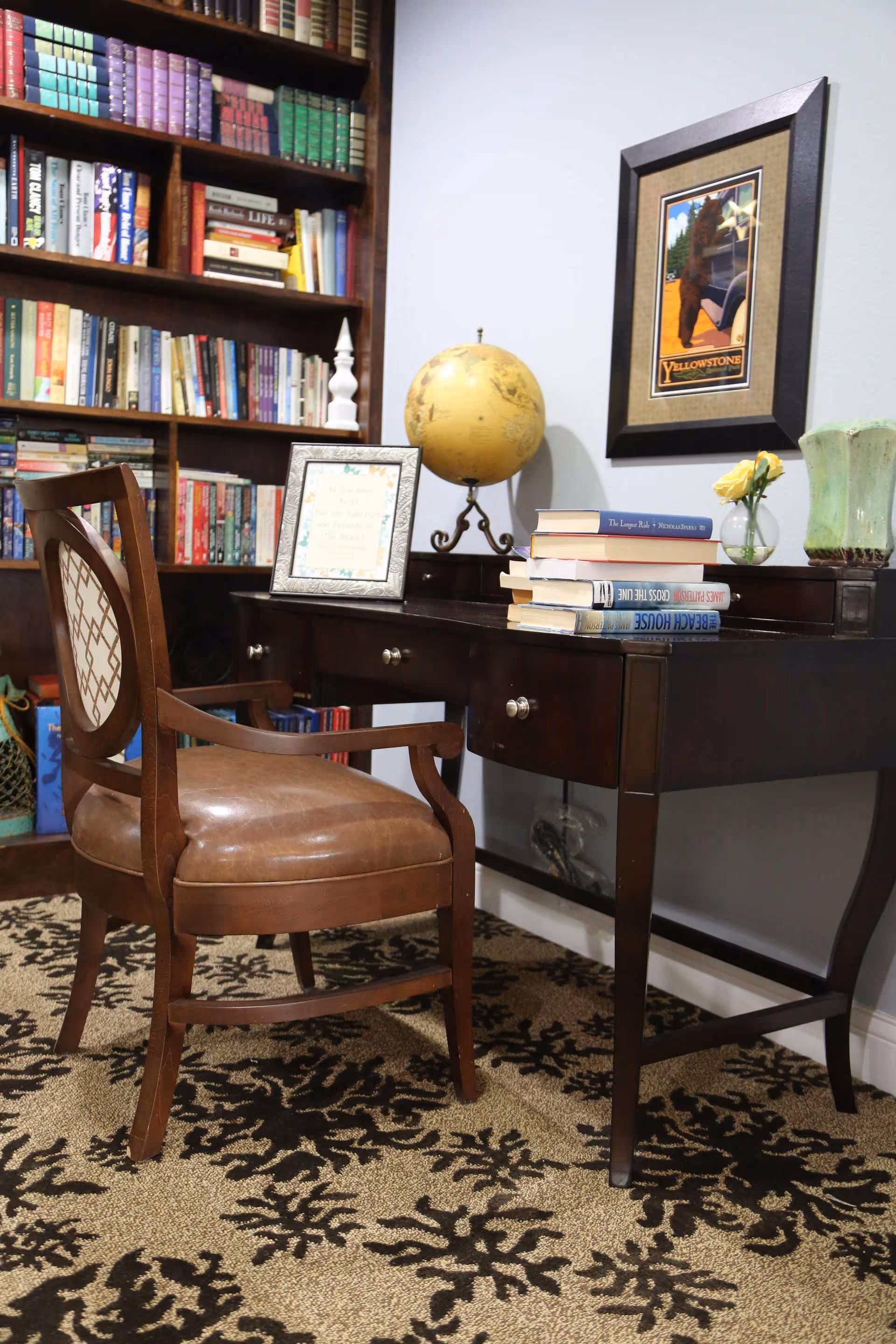 A cozy reading or study area featuring a wooden chair with a cushioned seat and backrest, a dark wooden desk with drawers, a globe, a stack of books, a framed picture, a small vase with yellow flowers, and a green decorative vase. Behind the desk is a bookshelf filled with various books, and a framed Yellowstone poster hangs on the wall. The floor is covered with a patterned carpet.