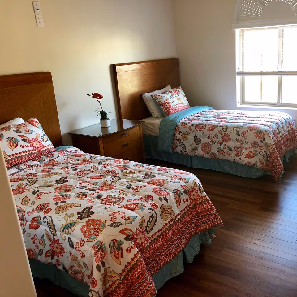 A bedroom with two single beds featuring wooden headboards and colorful floral-patterned bedding. A wooden nightstand with a small potted plant is placed between the beds. A window with blinds allows natural light into the room, and the floor is made of wood.