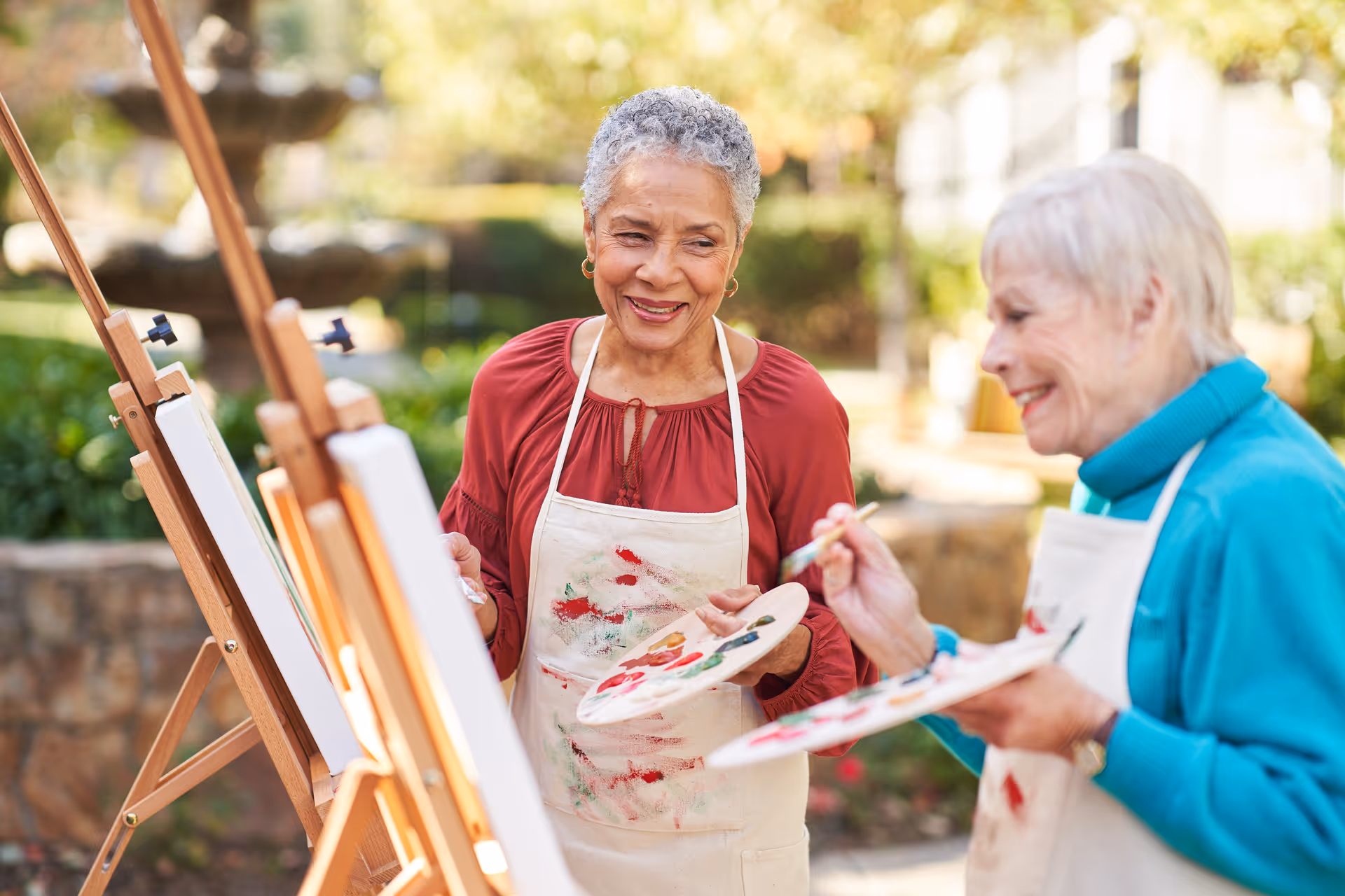 Two elderly women painting on canvases set on easels outdoors, both wearing aprons and smiling while engaging in the activity in a garden-like setting.