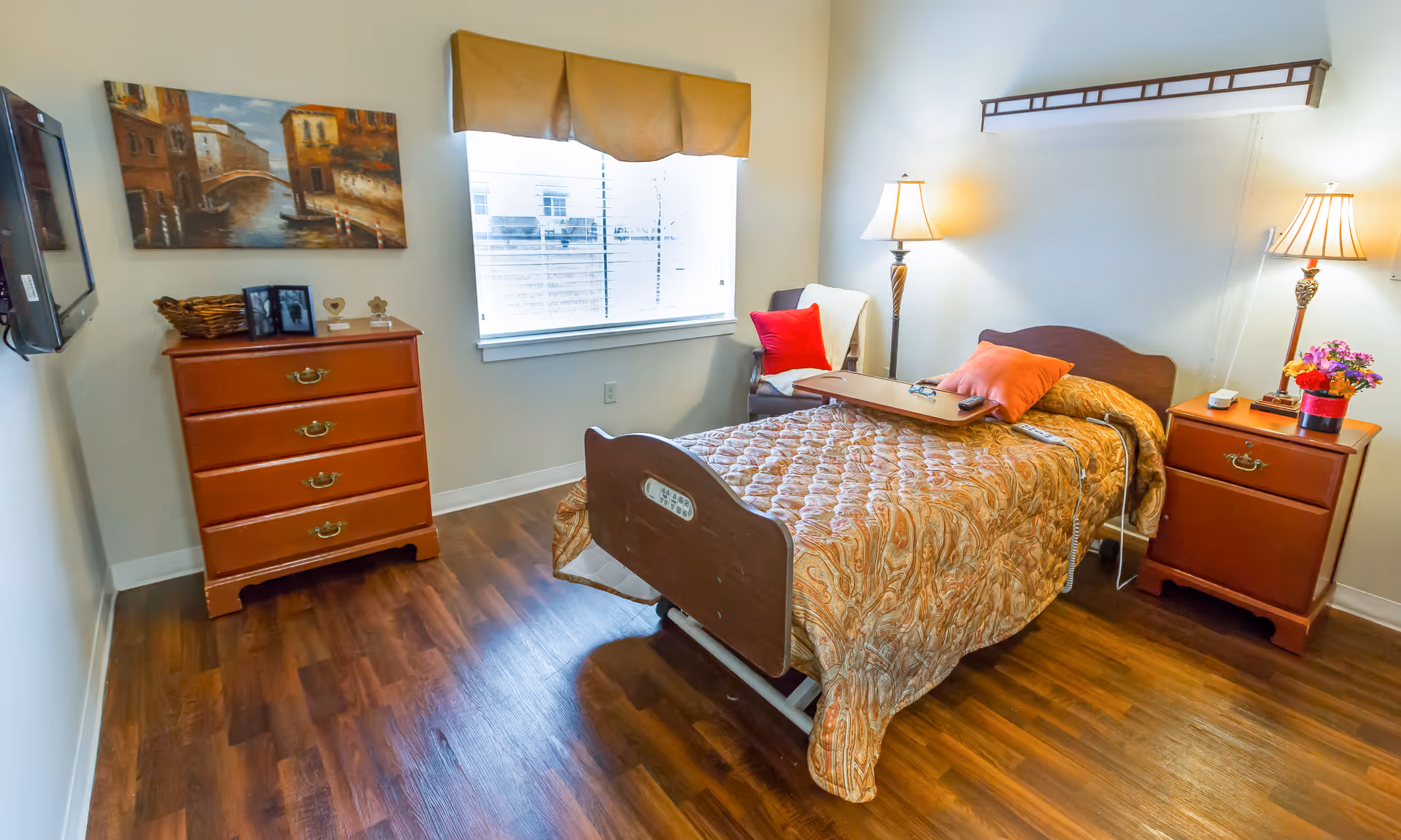 A well-lit bedroom in a senior living facility featuring a single bed with a patterned bedspread, a wooden headboard, and a tray table on top. There are two bedside tables with lamps and a small flower arrangement on one. A wooden dresser with four drawers is against the opposite wall, with a basket and framed photos on top. A window with a valance lets in natural light, and a comfortable chair with a red pillow is placed near the bed. The room has wood flooring and neutral-colored walls.
