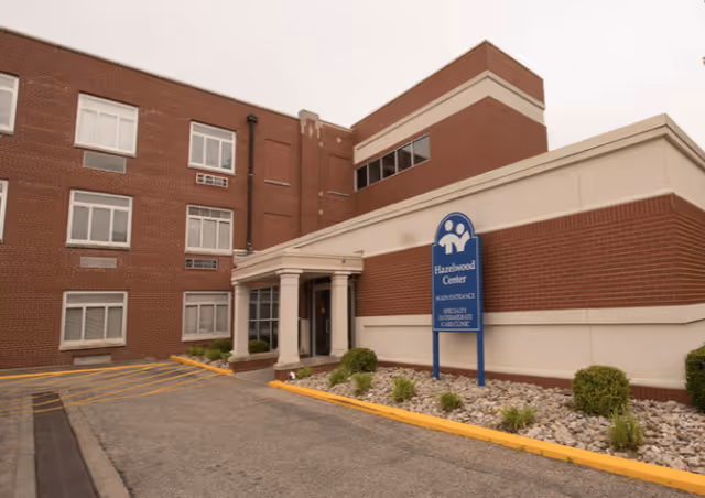 Exterior view of Hazelwood Center, a multi-story brick building with a beige and red facade. There is a blue sign in front of the building that reads 'Hazelwood Center, Skilled Nursing, Specialty Intermediate Care Unit.' The entrance has a small covered porch with columns, and there are small shrubs and rocks in the landscaped area beside the sidewalk.