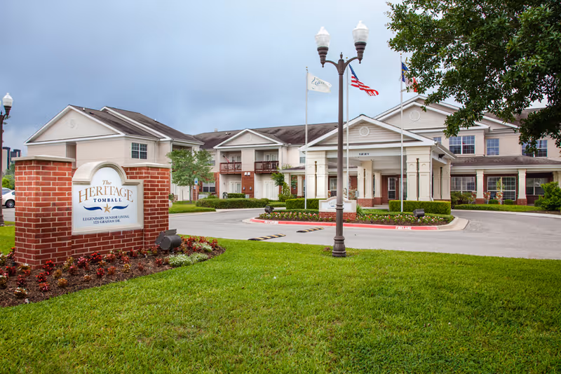Front exterior of The Heritage Tomball senior living facility showing the entrance, flagpoles, and a brick sign on a landscaped lawn.