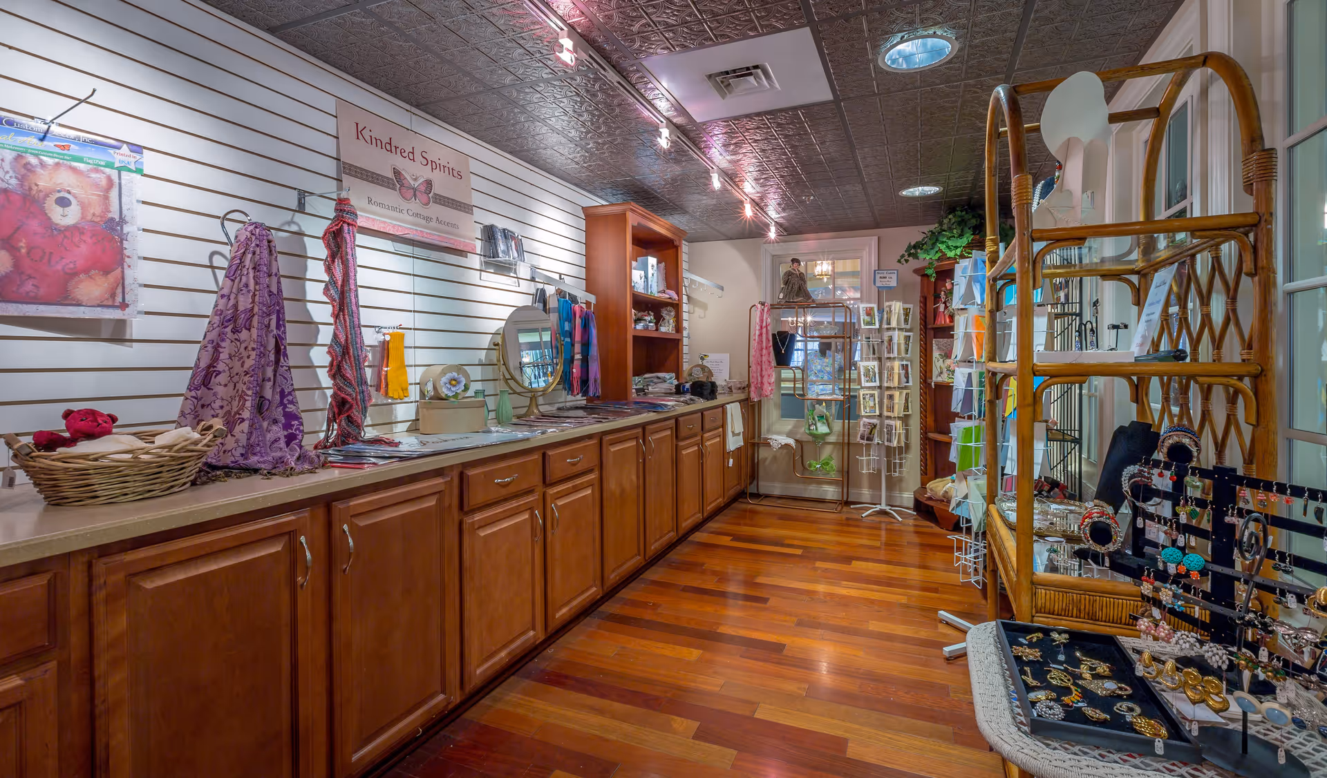 Interior view of a small gift shop area with wooden cabinets along the left wall displaying scarves, jewelry, and decorative items. There are shelves and racks filled with various accessories and gift items. The floor is wooden, and the ceiling has a decorative metal pattern with track lighting.