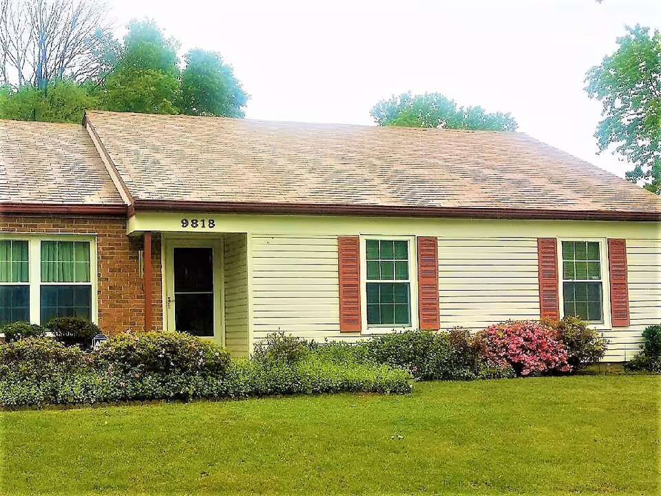 Single-story residential building with a brick and siding exterior, three windows with red shutters, a front door under a small covered porch, surrounded by green grass and bushes with some pink flowers.