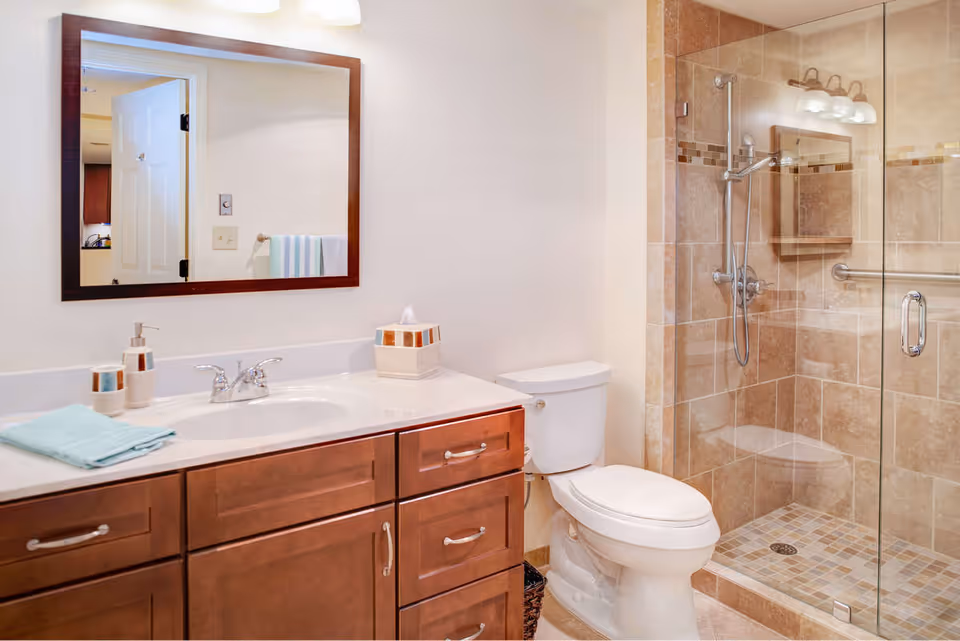 A clean and modern bathroom featuring a wooden vanity with a white countertop, a sink with a chrome faucet, a large mirror with a wooden frame, a white toilet, and a glass-enclosed shower with beige and brown tiles and a built-in bench.