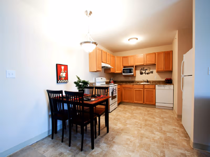 A kitchen and dining area with light wood cabinets, white appliances including a refrigerator, stove, microwave, and dishwasher. There is a small wooden dining table with four chairs, a plant centerpiece, and a red placemat. The walls are white, and the floor has beige tile. A decorative wall hanging with a chef and a sign that says 'Bon Appetit' are visible.