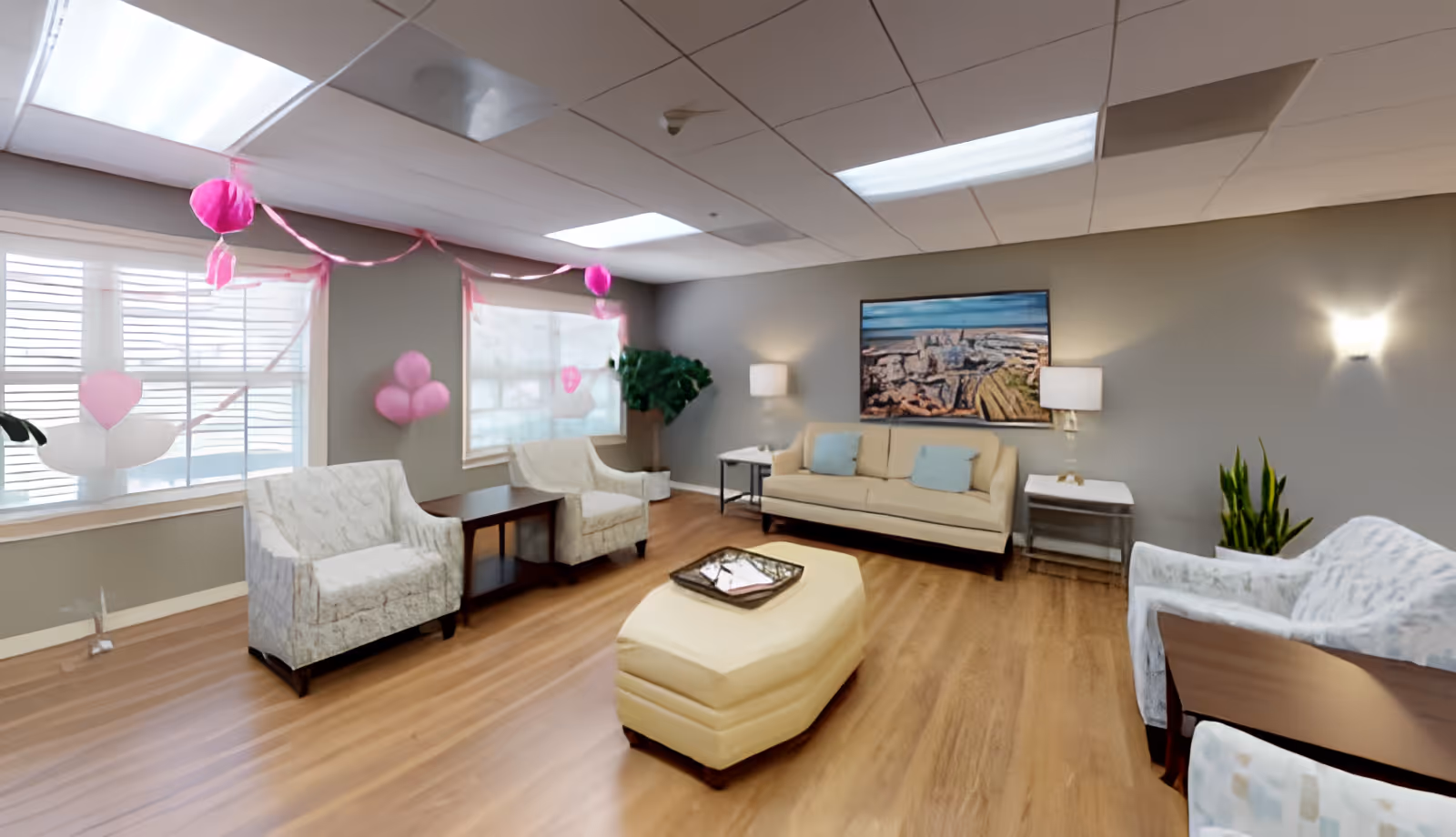 Well-lit senior living lounge with sofas, armchairs, an ottoman, wood floors, and pink balloon decorations.