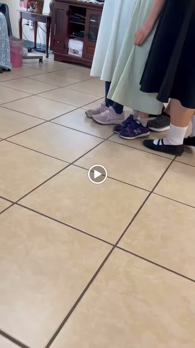 Partial view of four people standing indoors on a tiled floor, showing only their lower legs and feet. They are wearing different types of footwear including sneakers and flats, with some wearing socks. In the background, there is a wooden cabinet and a small table with an American flag decoration.