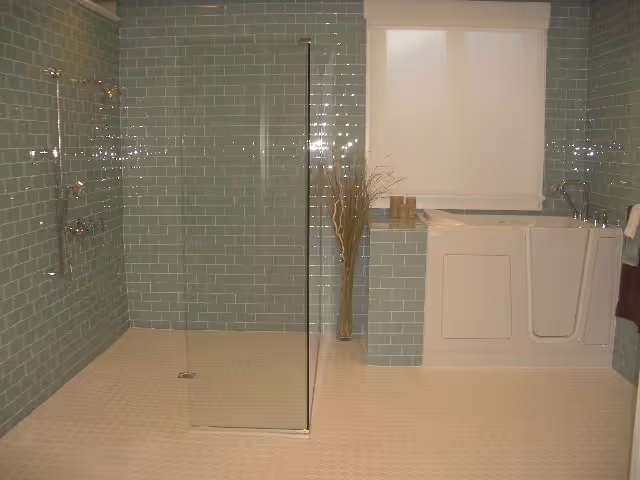 A bathroom with light green subway tile walls and beige hexagonal floor tiles. The room features a walk-in shower with a glass partition and a white walk-in bathtub with a faucet and a handrail. There is a window with a white blind above the bathtub and a tall vase with decorative branches next to it.