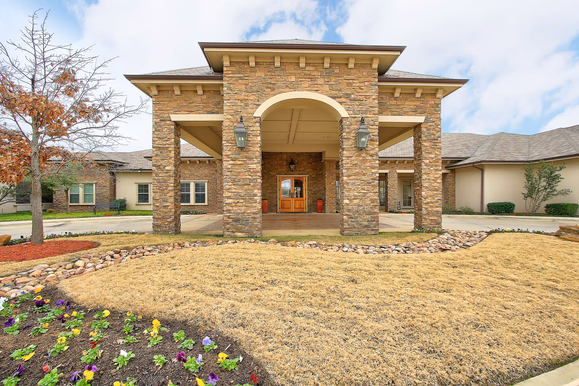 Front exterior view of a senior living facility with a stone entrance featuring large pillars, a covered entryway, and wooden double doors. The surrounding area includes a landscaped lawn with flower beds and a small tree.