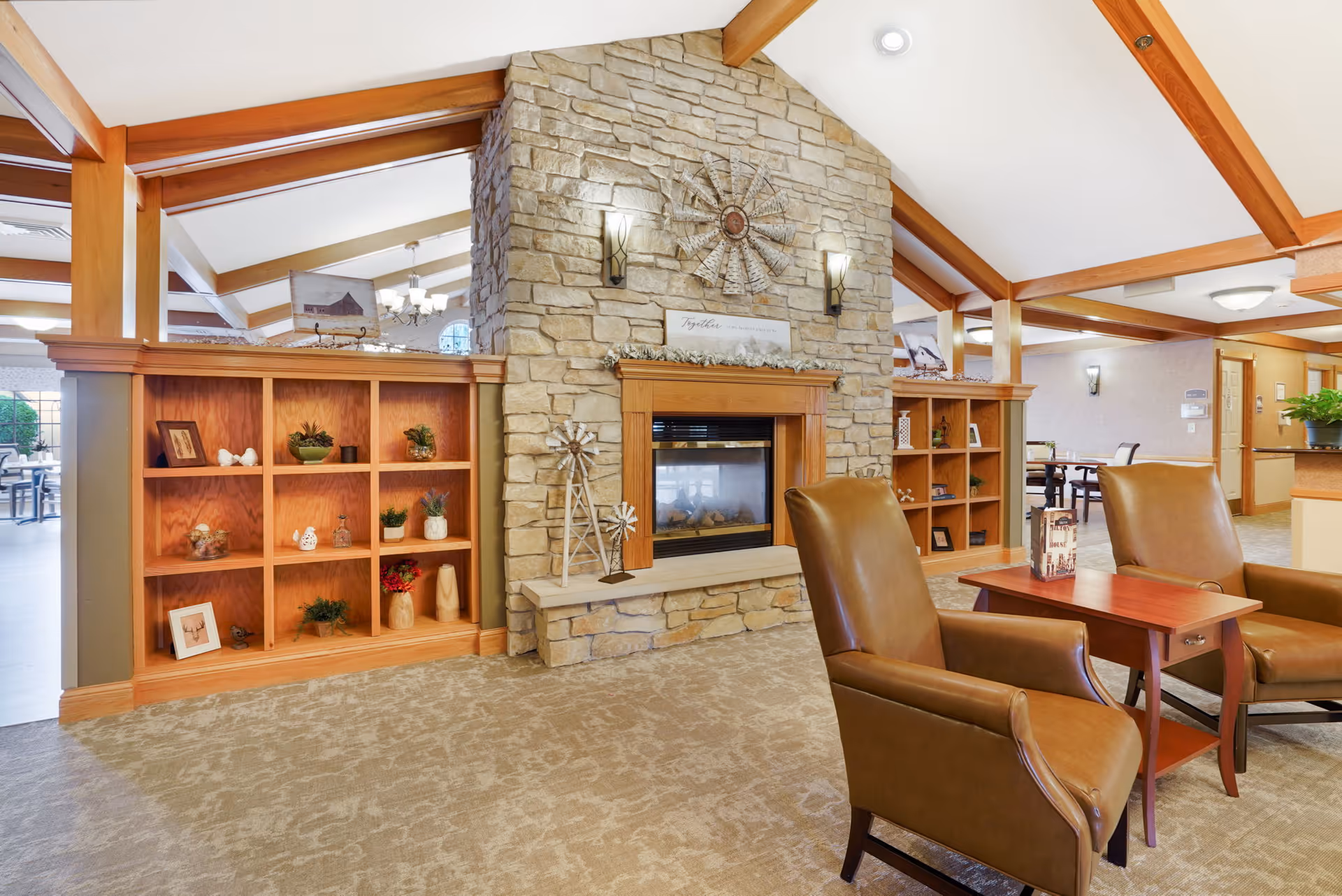 A cozy senior living facility common area featuring a stone fireplace with decorative windmill accents, wooden built-in shelves with plants and ornaments, two brown leather armchairs, and a small wooden table. The room has exposed wooden beams on a vaulted ceiling and carpeted flooring.