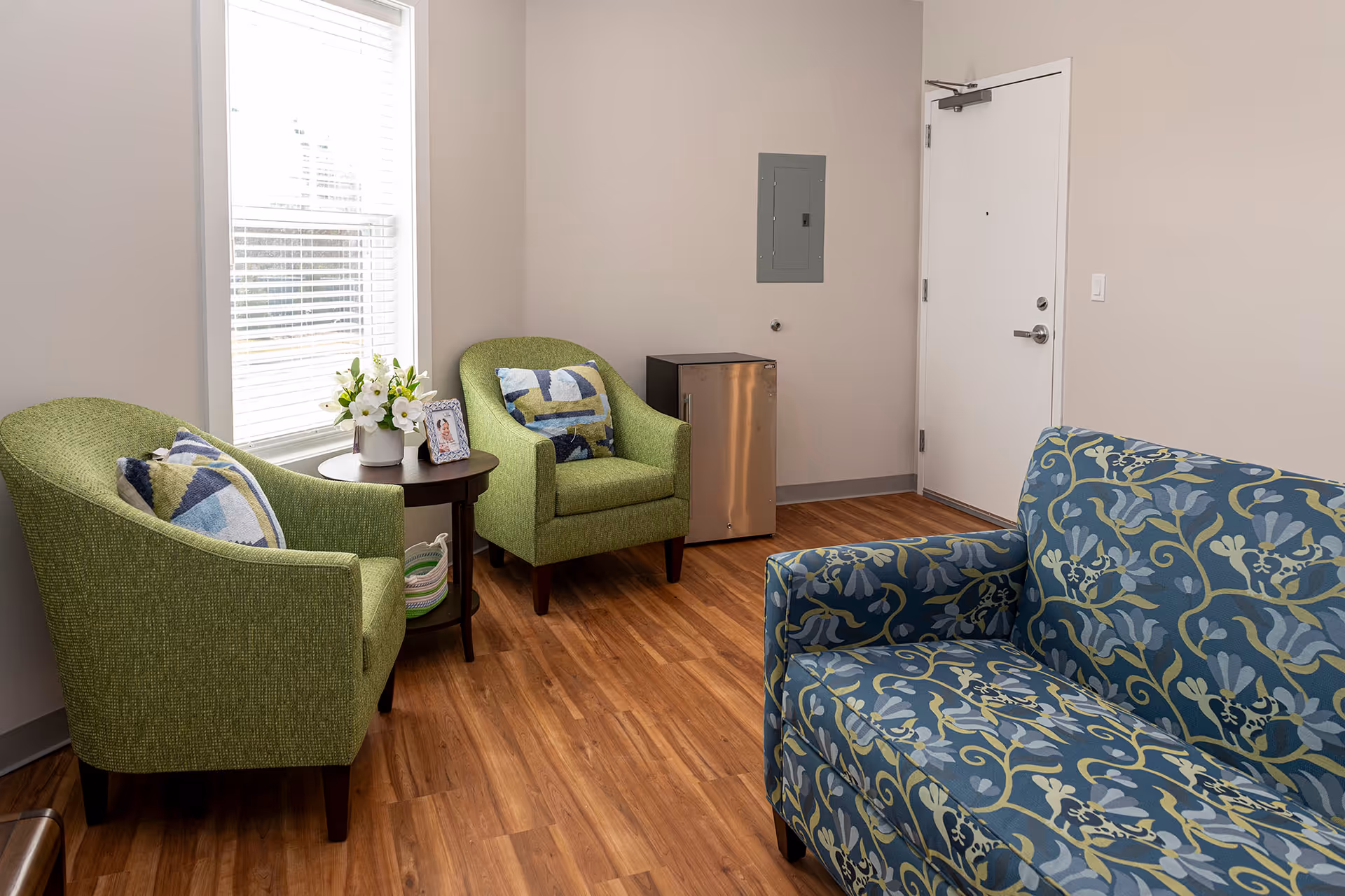 A small sitting area with two green armchairs and a blue patterned loveseat arranged around a round wooden side table with a vase of white flowers and a framed photo. The room has wood flooring, a window with blinds, a white door, and a small stainless steel mini fridge against the wall.