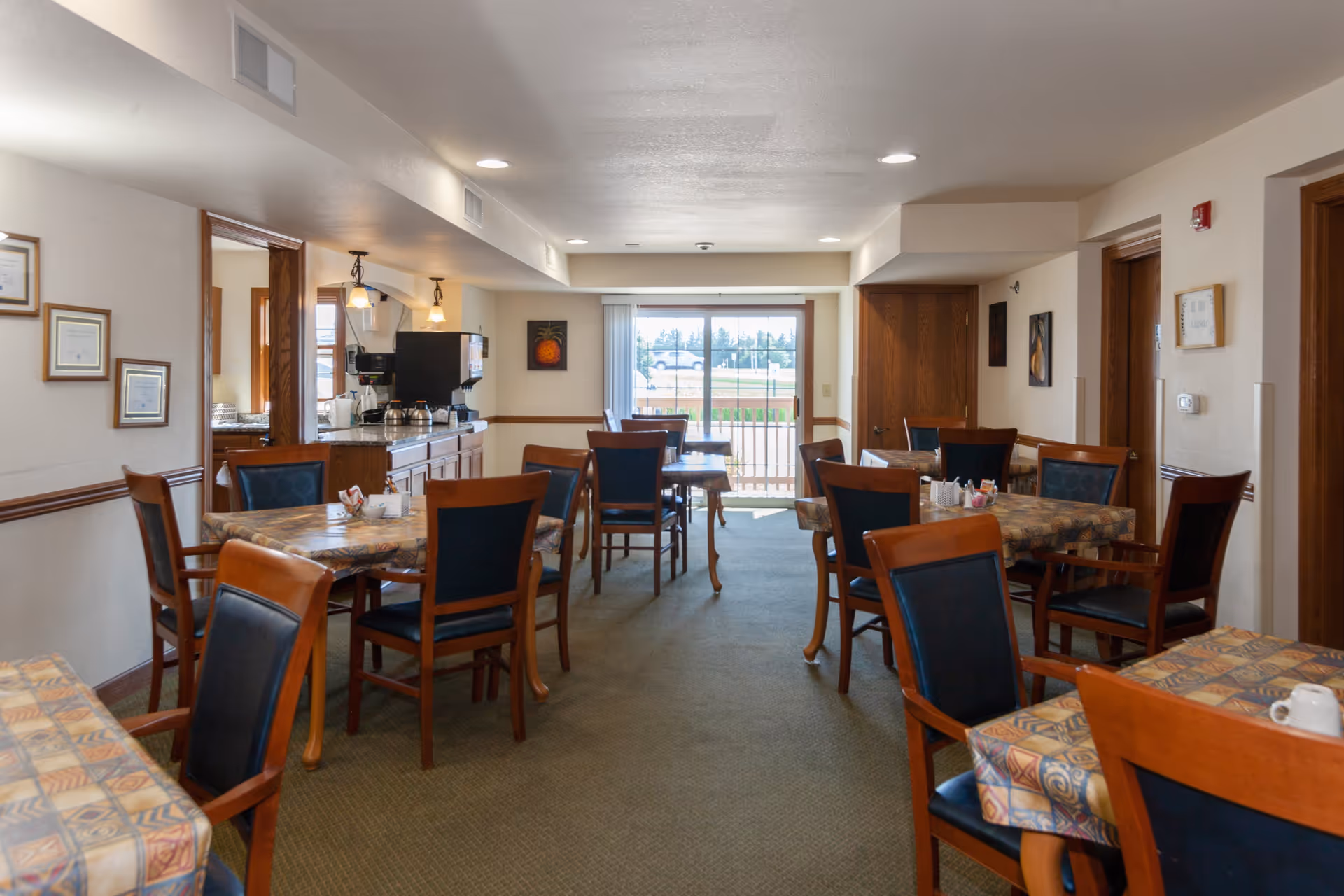 Dining room with multiple tables and chairs, patterned tablecloths, a coffee station on the left, and a sliding glass door at the far end.