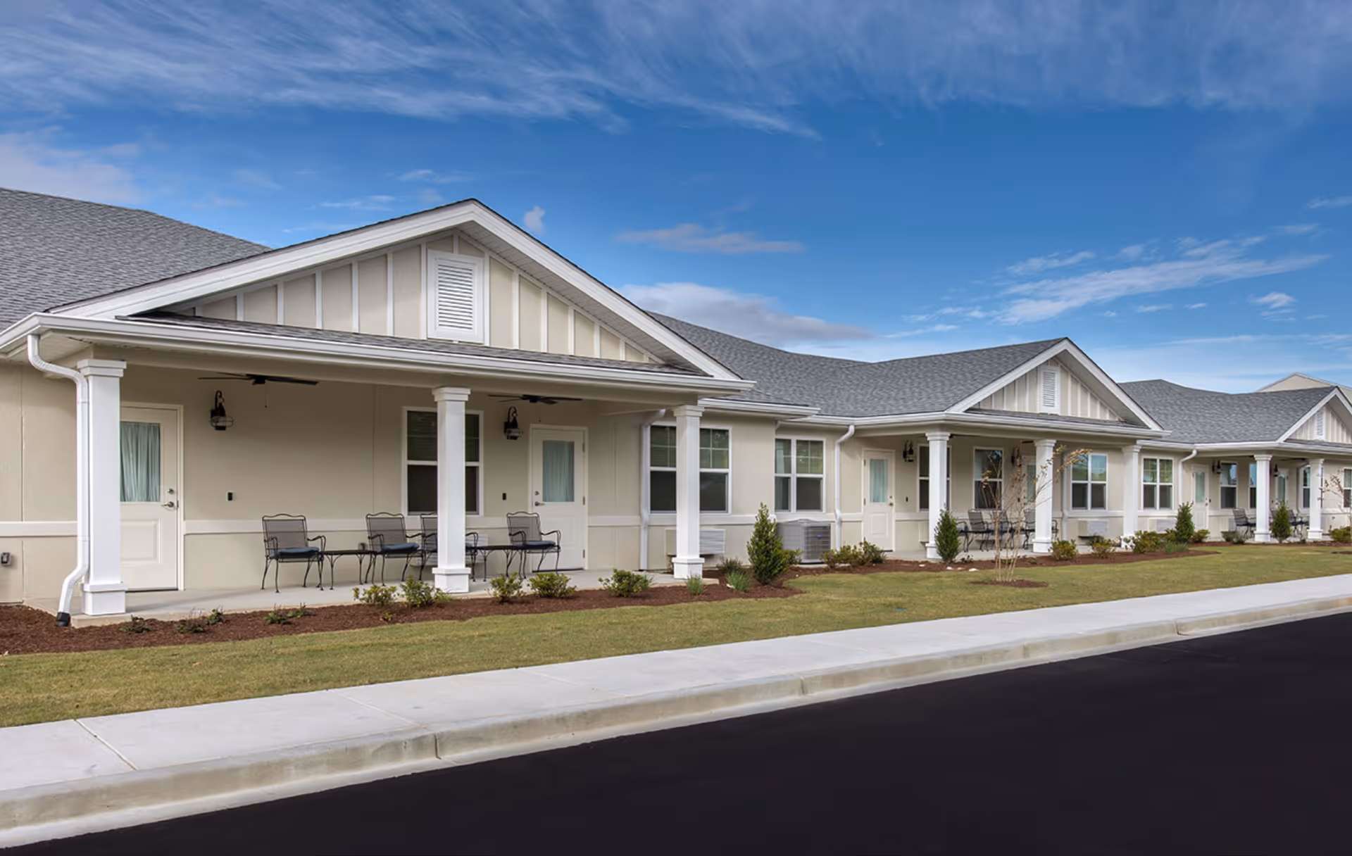 Single-story senior living building frontage with covered porches, outdoor seating, and a landscaped lawn under a blue sky.