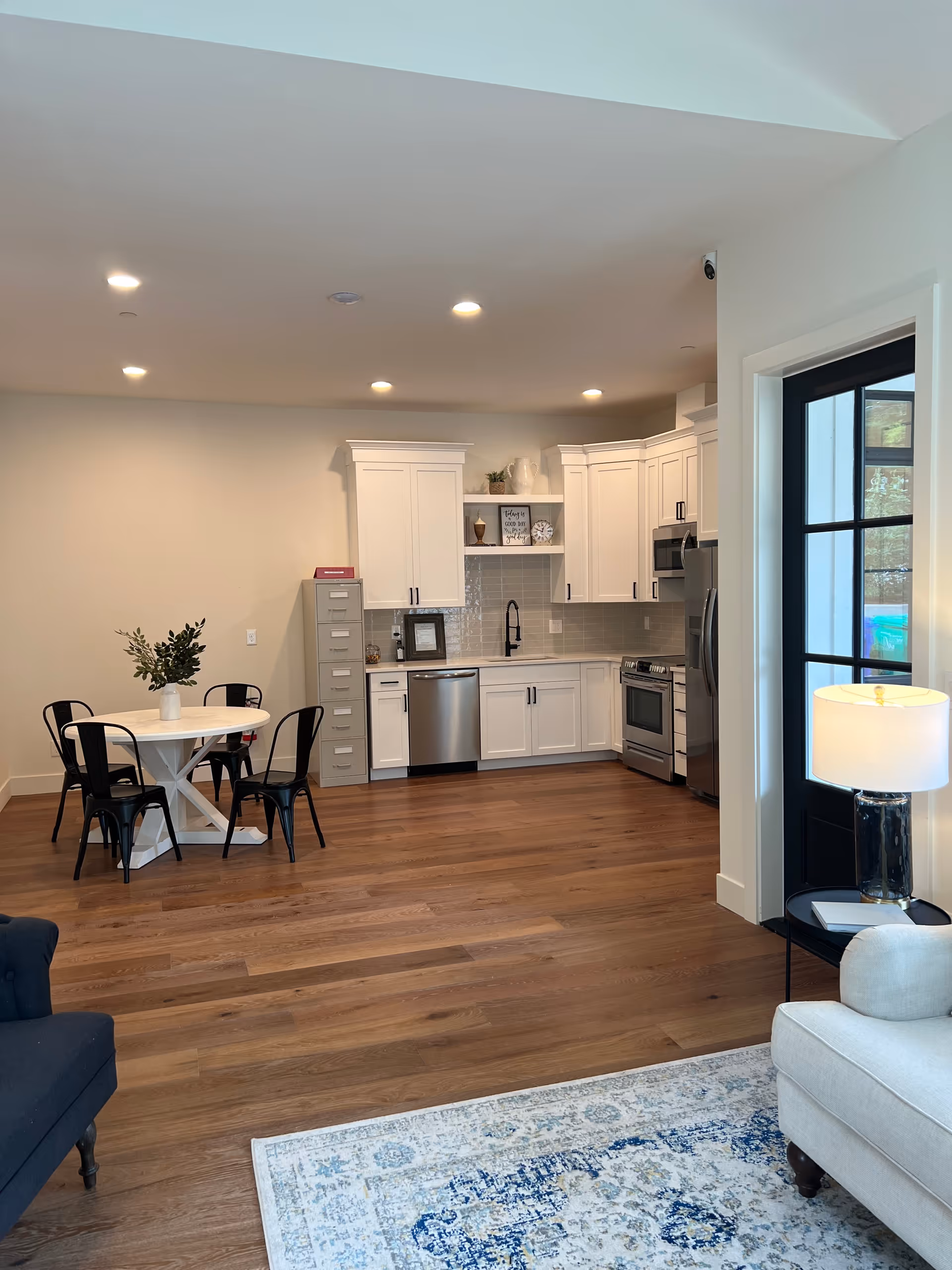 Open-plan living area with white kitchen cabinets, stainless steel appliances, a round dining table, and wood floors.