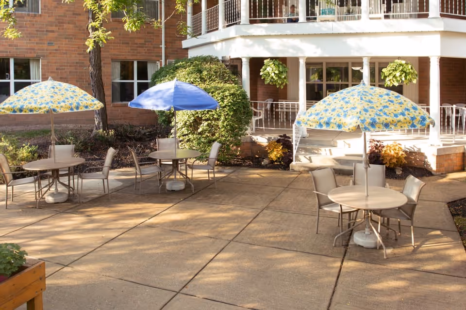Outdoor patio area with round tables and chairs, each table shaded by umbrellas in blue and sunflower patterns. The patio is adjacent to a brick building with windows and a covered porch with white railings and hanging plants.