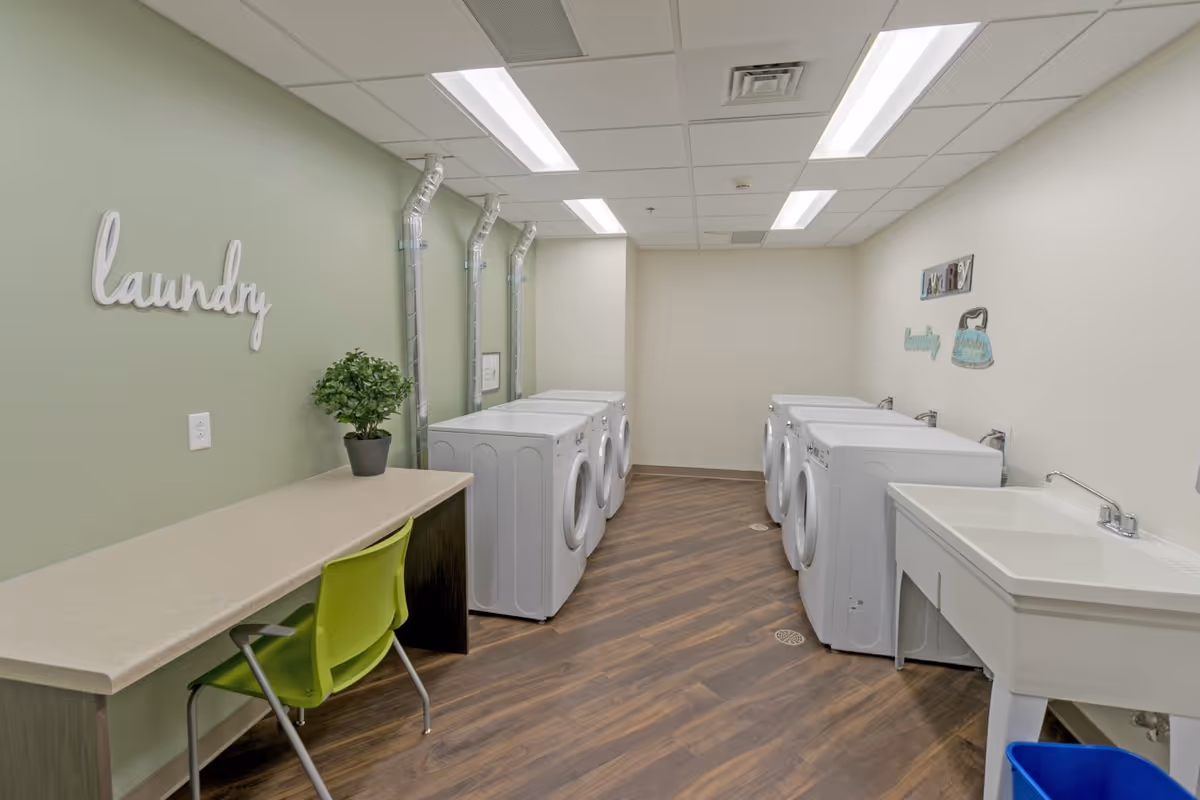 Bright laundry room with multiple front-loading washers, a utility sink, and a folding counter with a green chair.