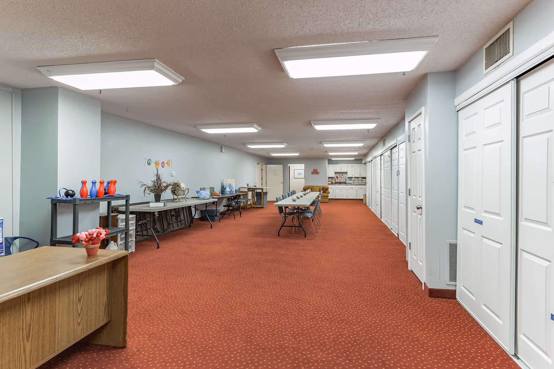 A long room with red carpet and light blue walls featuring a row of white folding doors on the right side. In the center, there is a long table with chairs arranged around it. The far end of the room has a small kitchen area with white cabinets and a brown couch. On the left side, there are folding tables with various items including a small bowling set and decorative objects. The ceiling has multiple rectangular fluorescent lights.