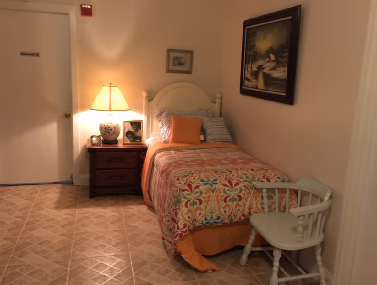 A small bedroom with a single bed covered in a colorful patterned bedspread and orange blanket. There is a wooden nightstand with a lamp and framed photos next to the bed. A white wooden chair is placed near the foot of the bed. The walls are light-colored with a framed painting hanging above the bed. The floor has a tiled pattern.