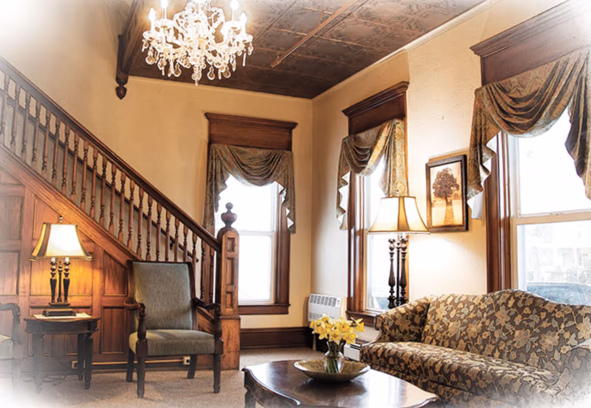 Cozy traditional living room with a patterned sofa, armchair, wooden staircase, chandelier, lamps, and draped windows.