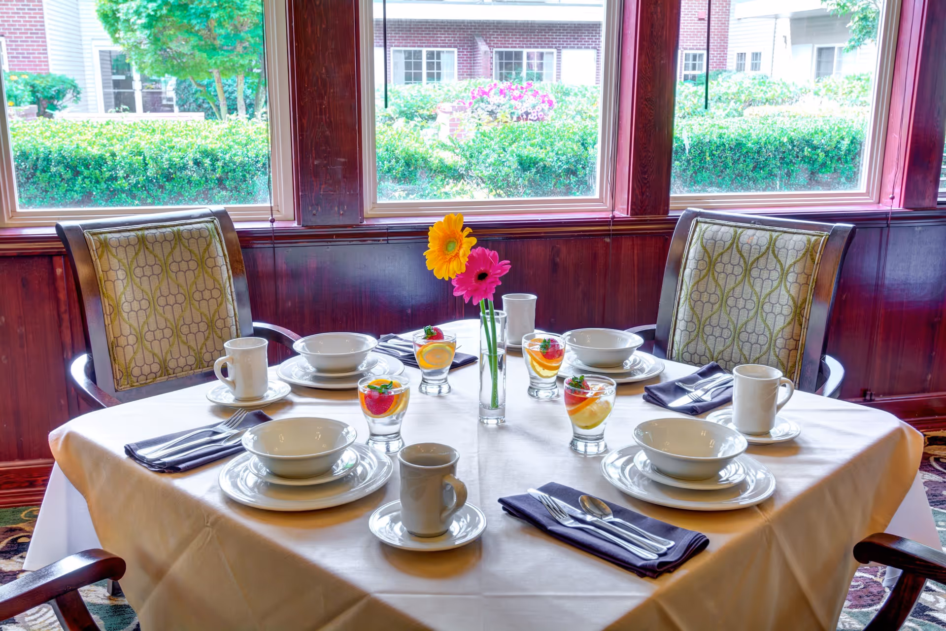 A dining table set for four with white tablecloth, white plates, bowls, cups, silverware on dark napkins, and glasses with fruit-infused water. A small vase with two colorful flowers is in the center. The table is near large windows showing greenery and a building outside.