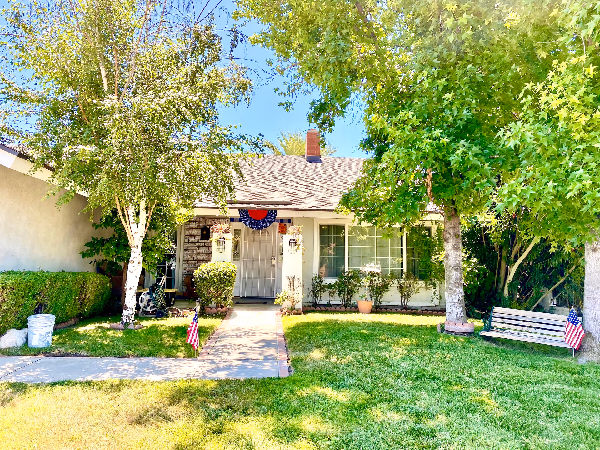 Front exterior view of a single-story house with a well-maintained lawn, two trees, a bench with an American flag, and a walkway leading to the front door decorated with a red, white, and blue fan-shaped banner.