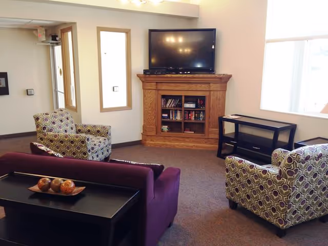A cozy living room area with a purple sofa and two patterned armchairs facing a wooden TV stand with a flat-screen television on top. The TV stand has shelves with books and other items. There is a small black table next to the window, and the room has beige walls and carpeted flooring.