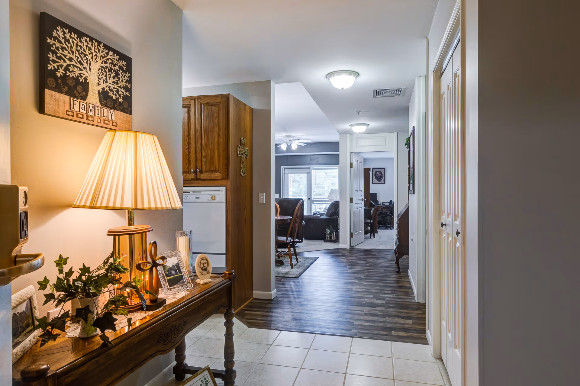 Interior view of a senior living facility hallway with a wooden console table on the left holding a lamp, framed photos, a decorative cross, and a plant. The hallway leads to a dining area with wooden chairs and a living room with large windows and a ceiling fan. The floor transitions from tiled near the entrance to wood laminate further inside.