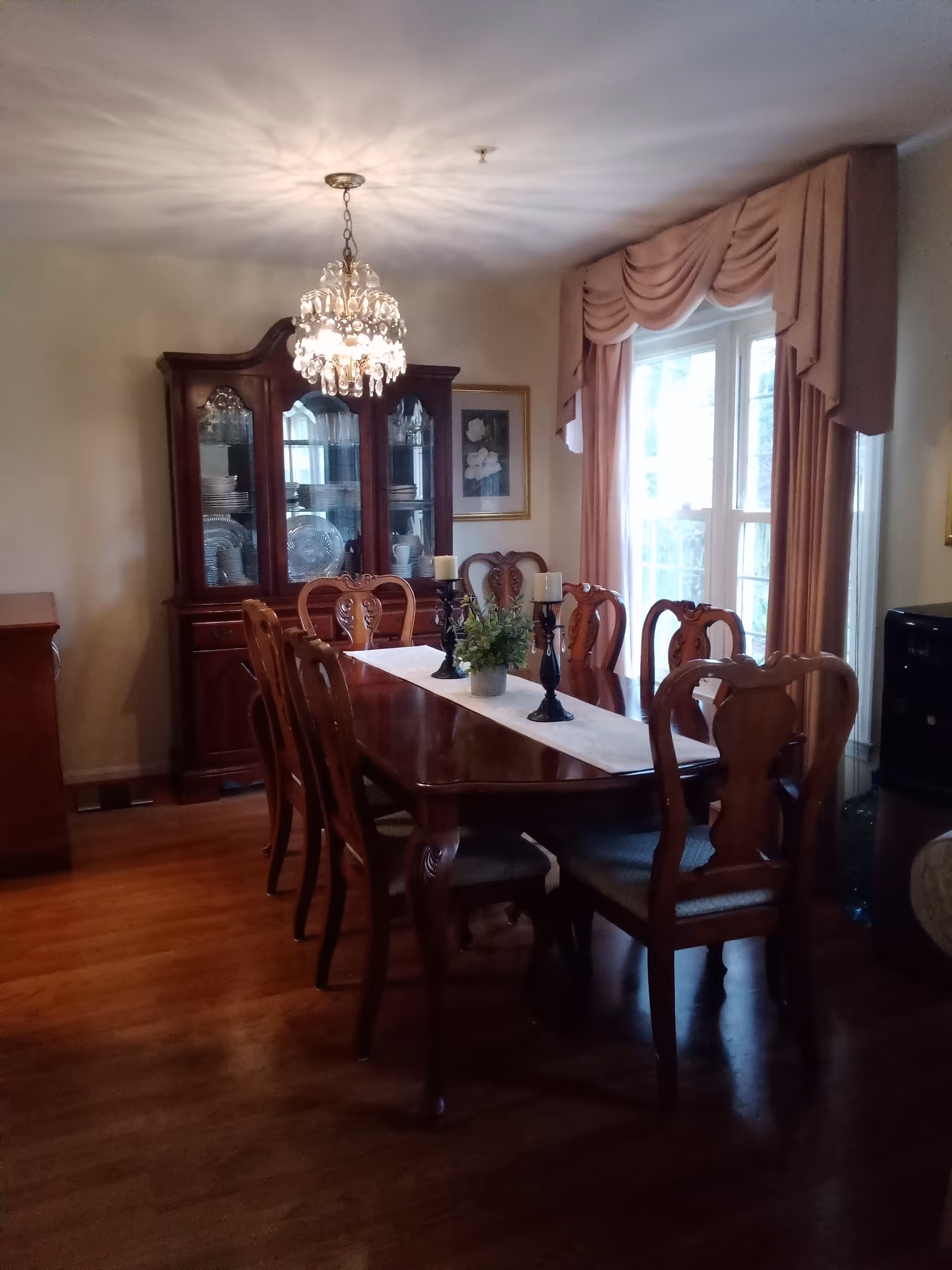 A traditional dining room with a long wooden dining table surrounded by eight matching wooden chairs. The table is decorated with a white runner, a small potted plant, and three black candle holders with white candles. A crystal chandelier hangs above the table, casting light patterns on the ceiling. In the background, there is a wooden china cabinet displaying plates and glassware. The room has hardwood flooring, a large window with pink drapes, and framed artwork on the walls.