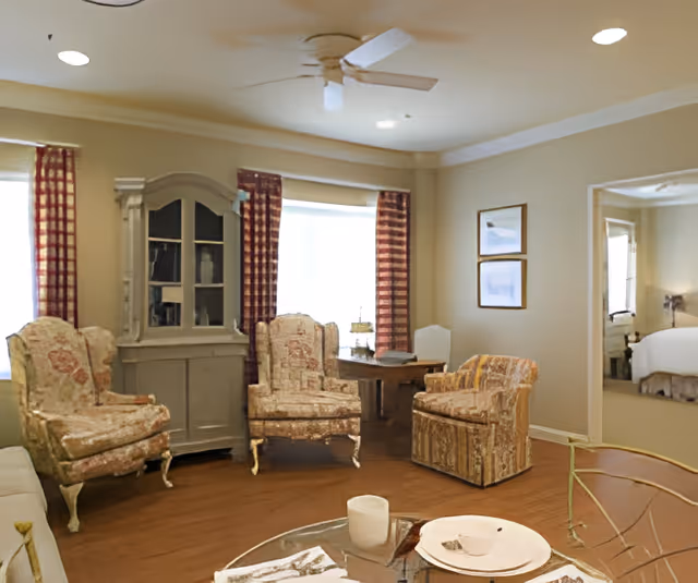 A cozy living room in a senior community featuring three upholstered armchairs with floral and patterned designs, a glass coffee table with a plate and cup, a wooden cabinet with glass doors, two windows with red checkered curtains, a ceiling fan, and a glimpse into a bedroom through an open doorway.
