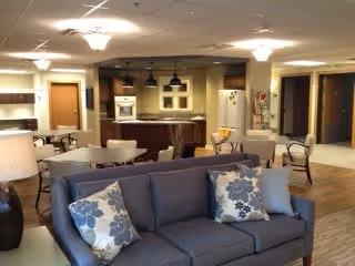 A spacious and well-lit common area featuring a modern kitchen with dark wood cabinets, a white refrigerator, and a microwave. In the foreground, there is a gray sofa with floral patterned pillows. Several white round tables with chairs are arranged around the room, and there are multiple doorways leading to other rooms. The ceiling has recessed lighting and decorative light fixtures.