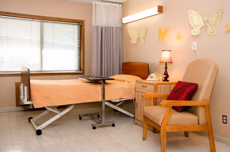A senior living facility room with a hospital-style bed covered in a peach blanket, a wooden nightstand with a lamp and a telephone, a beige armchair with a red pillow, and butterfly wall decorations. There is a window with blinds and a privacy curtain partially drawn.