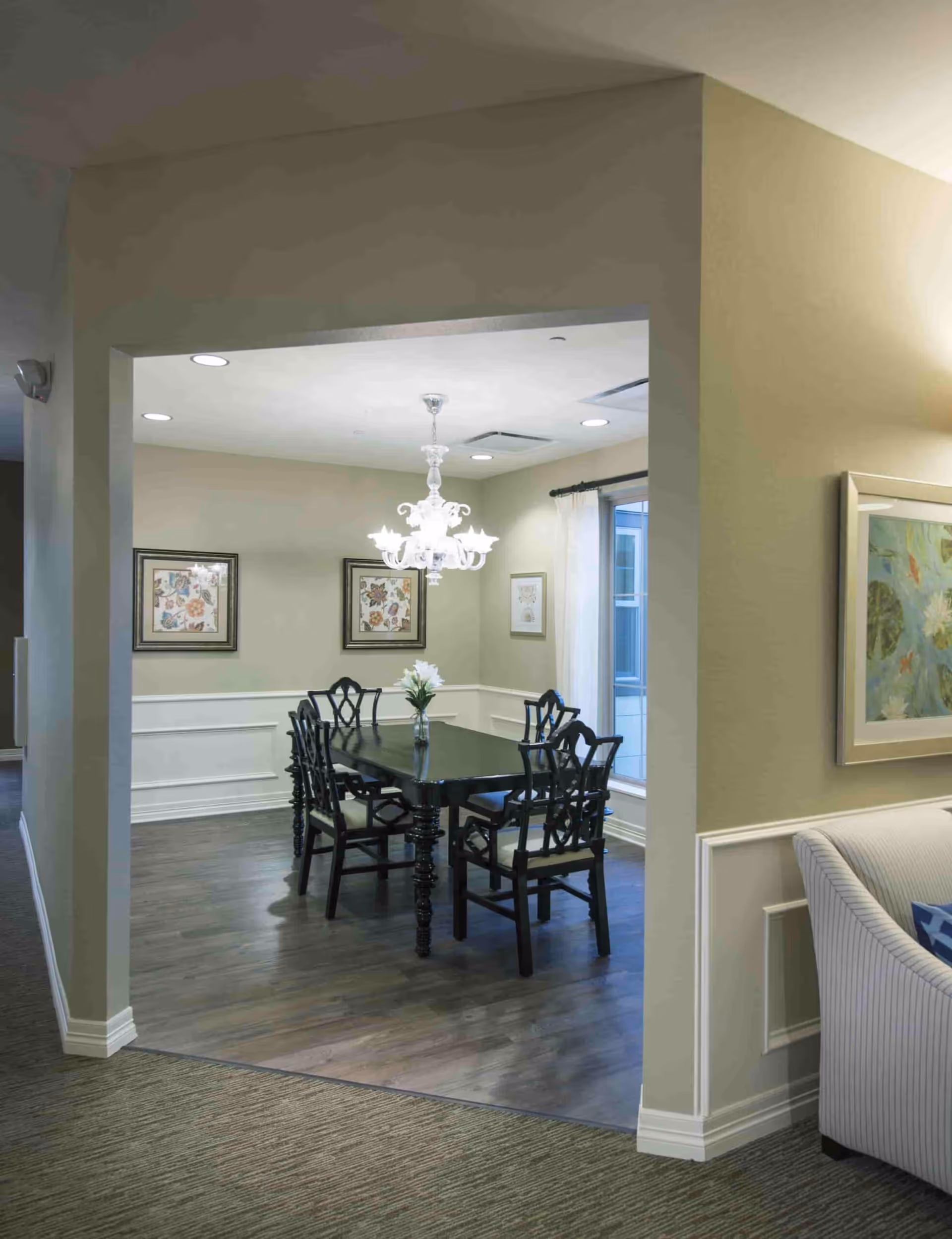A dining room with a dark wooden table and six matching chairs. A vase with white flowers is placed in the center of the table. The room has light-colored walls with white wainscoting, two framed floral artworks on one wall, and a window with white curtains. A white chandelier hangs above the table. Adjacent to the dining room is a living area with a striped armchair and a framed painting on the wall.