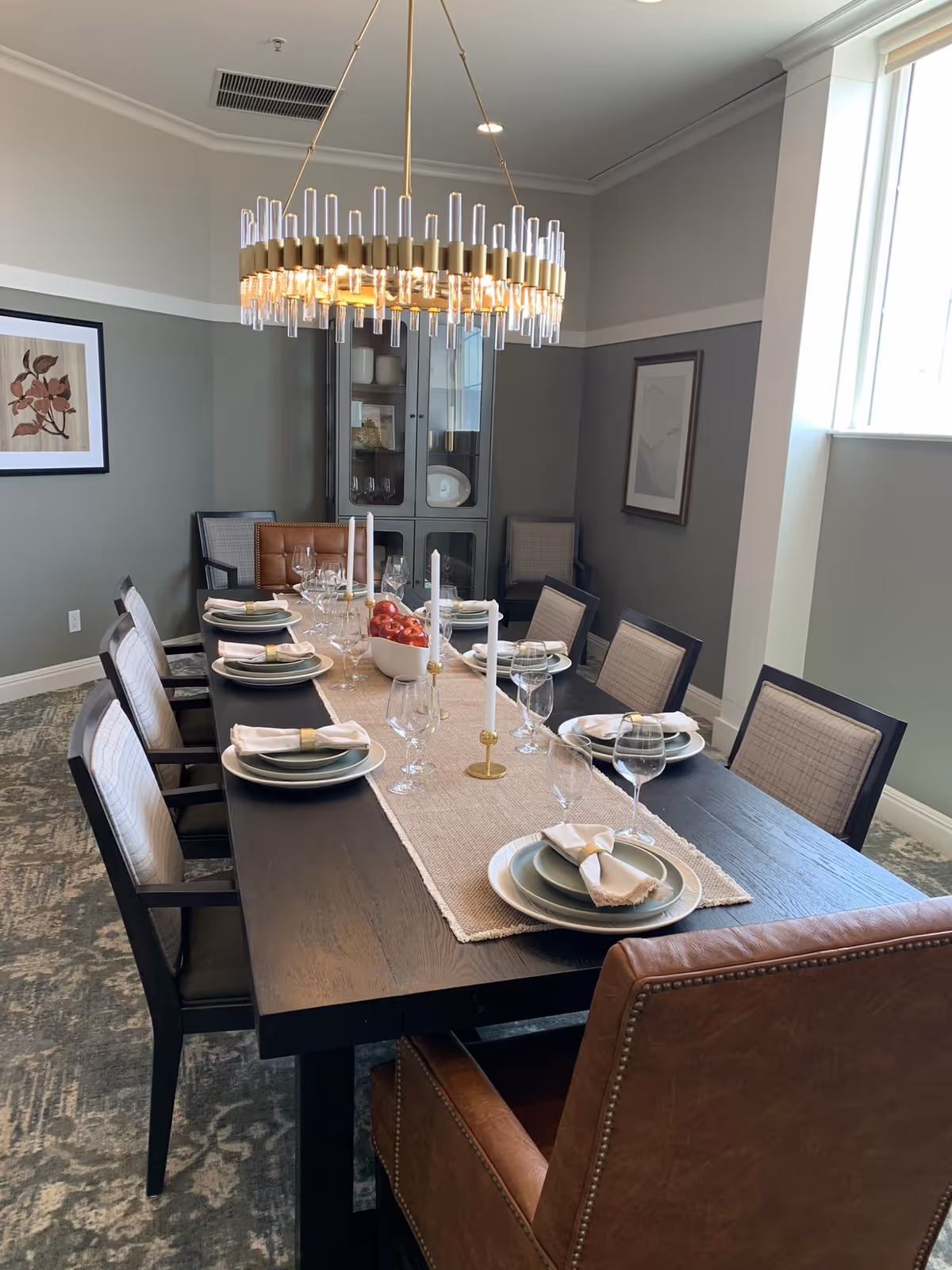 A dining room with a long dark wooden table set for eight people. The table is decorated with a beige runner, white plates with napkins, wine glasses, and three white candles in gold holders. A bowl of red apples is placed in the center. The room has gray walls with white trim, two framed pictures, a glass-front cabinet, and a modern chandelier hanging above the table. There is a large window letting in natural light.
