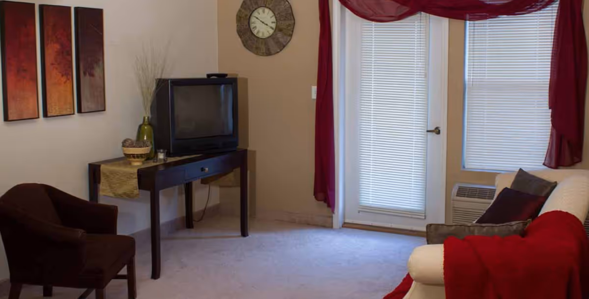 A cozy living room with a white couch draped with a red blanket and several pillows, a dark brown armchair, a wooden table with a vintage TV on top, decorative items including a green vase and a bowl, a wall clock, and three-panel artwork on the wall. There is a door and window with blinds and red curtains.